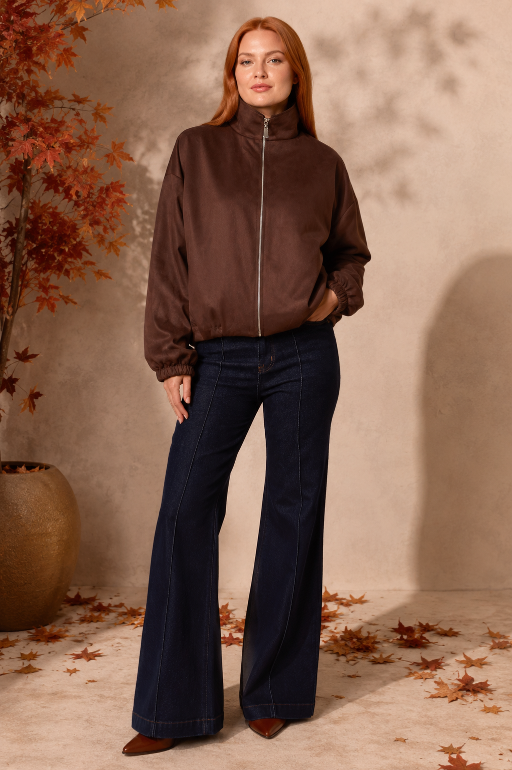 A woman stands in a studio setting wearing a brown zip-front jacket and dark navy wide-leg jeans, with autumn leaves and a potted plant in the background.