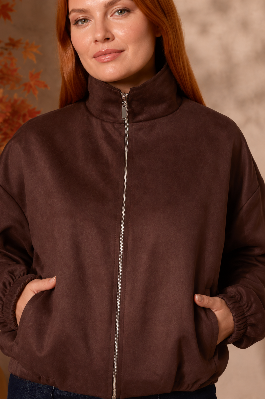 A woman wearing a dark brown zip-up jacket with a high collar and elasticized cuffs, standing against a warm-toned textured background.