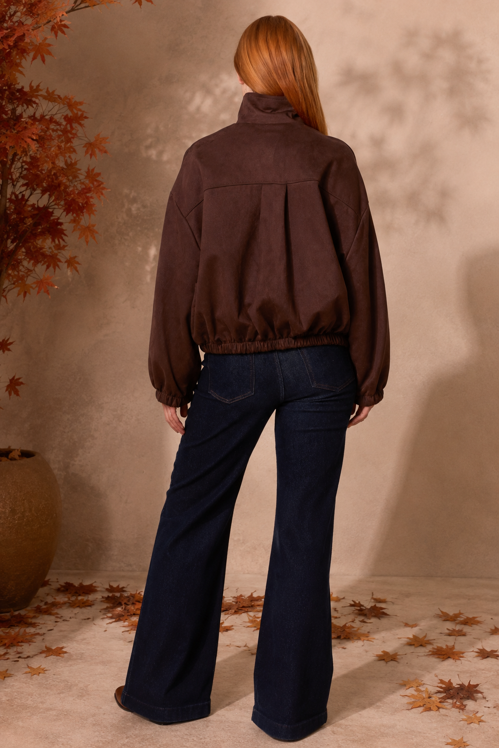 A woman shown from behind wearing a dark brown cropped jacket and dark blue wide-leg jeans, standing in a styled indoor setting with a textured beige wall, scattered autumn leaves, and a potted plant.