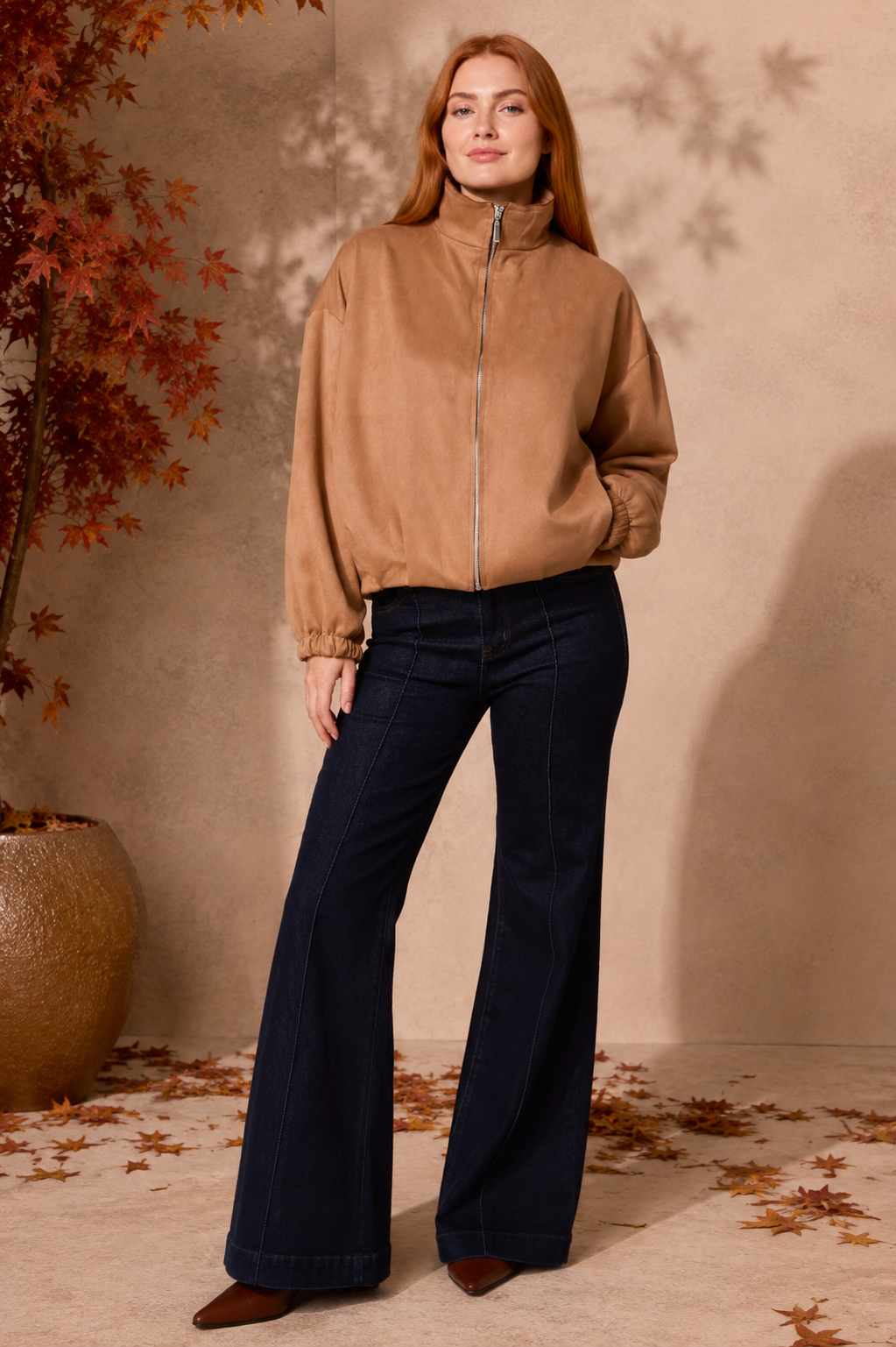 A woman stands in a styled indoor setting wearing a tan zip-front jacket with a high mock neck, dark navy wide-leg jeans, and brown pointed shoes. The background features a textured beige wall with leaf shadows, a potted autumn tree, and scattered fallen leaves on the floor.