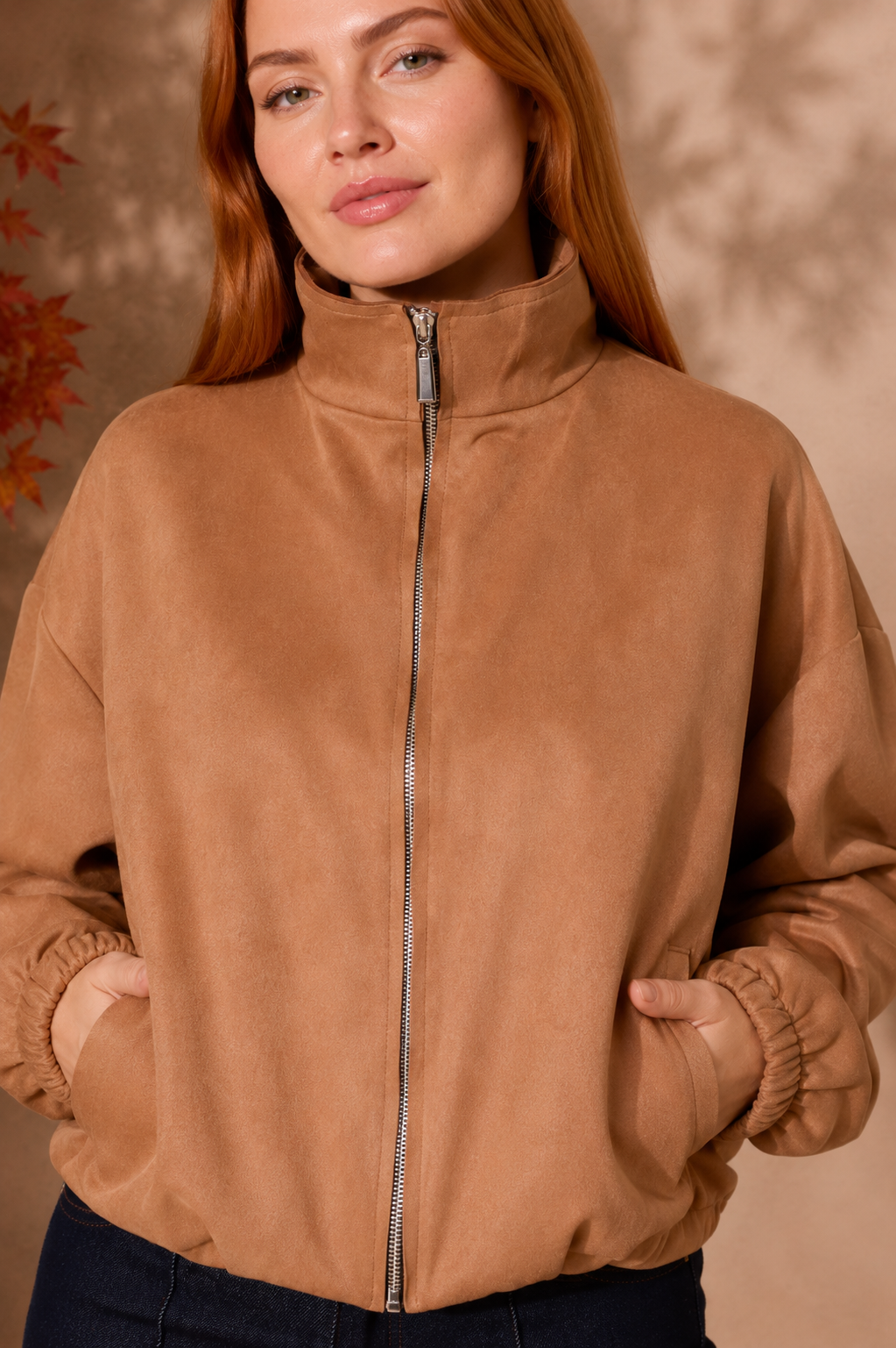 A woman wearing a camel brown faux-suede zip-up jacket with a high stand collar and elasticized cuffs, paired with dark blue jeans.