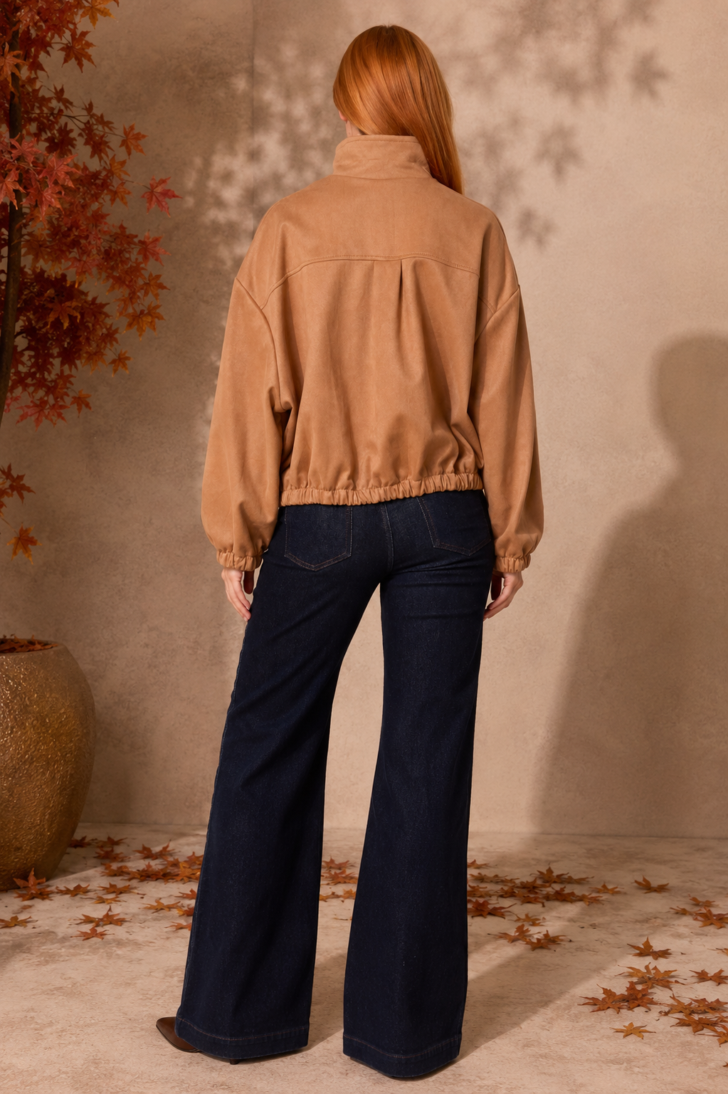 A woman with long red hair is shown from behind wearing a tan cropped jacket and dark blue wide-leg jeans, standing in a styled indoor setting with a textured beige wall, scattered autumn leaves, and a potted plant.