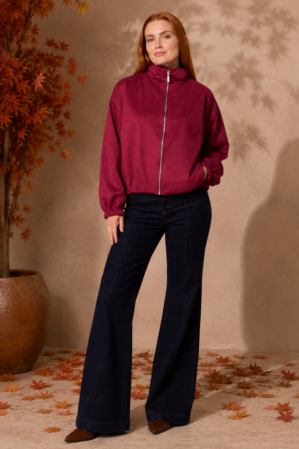 A woman stands in a studio setting wearing a burgundy zip-front jacket with a high collar and dark navy wide-leg jeans. The background is a warm beige wall with leaf shadows, a potted autumn tree on the left, and scattered orange leaves on the floor.
