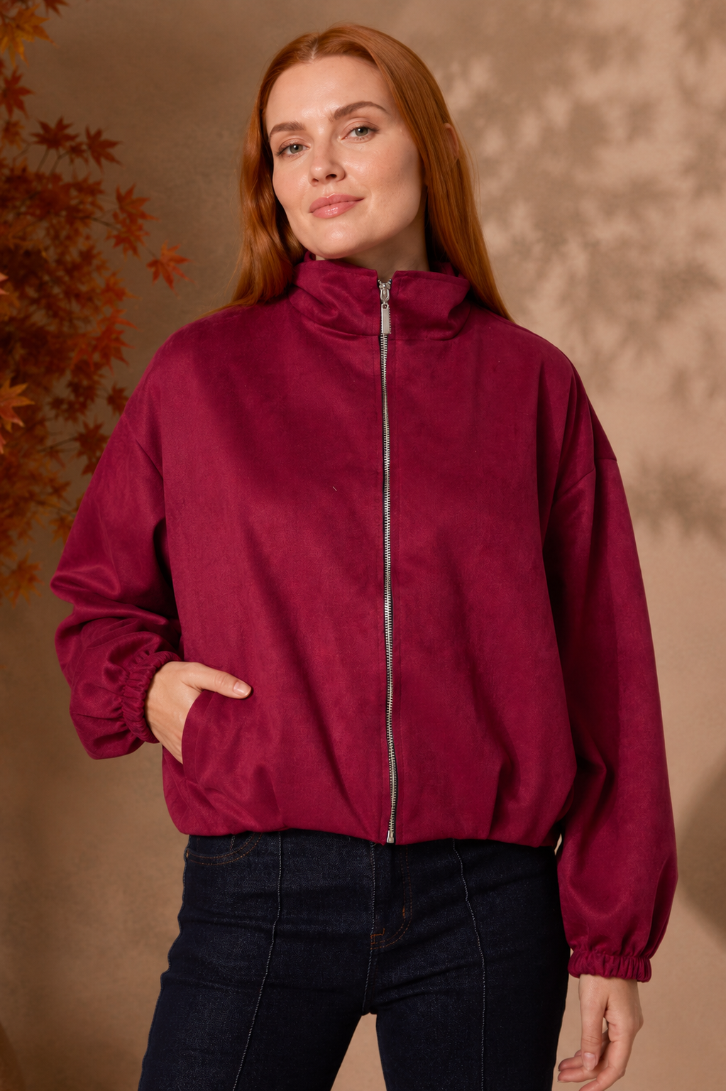 A woman with red hair wearing a cropped, long-sleeve zip-up jacket in deep burgundy with dark blue jeans, standing against a warm brown textured background.