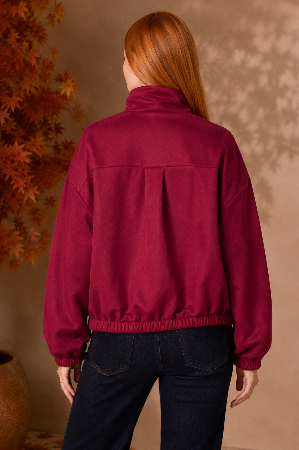 A woman with long red hair is shown from the back wearing a dark magenta fleece jacket and dark blue jeans, standing against a warm beige wall with autumn leaves on the left.